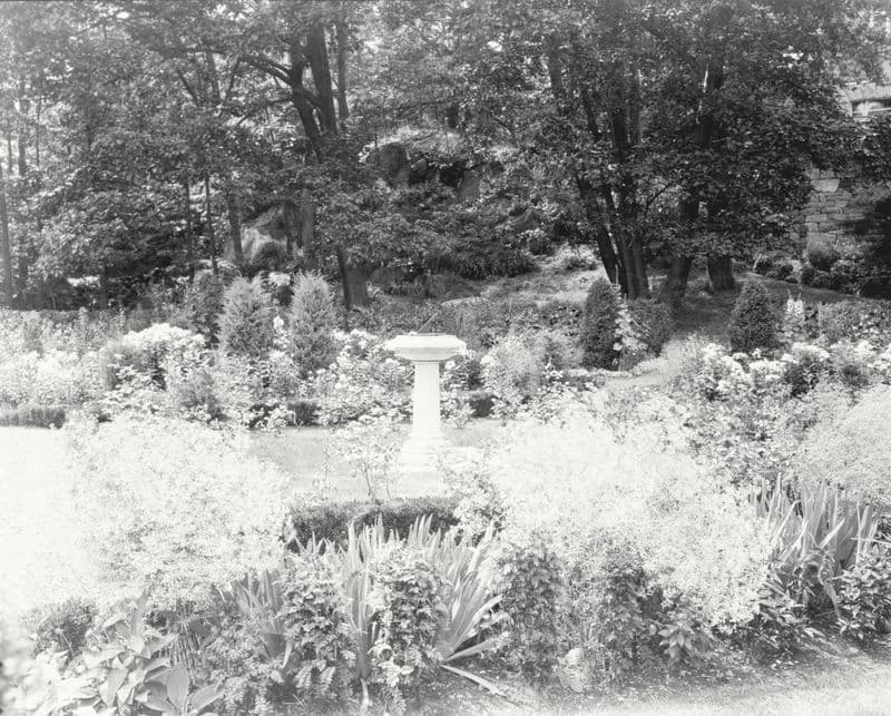 A pedestal sundial among flower beds and clipped evergreens in a New England garden.